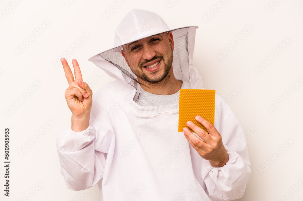 Hispanic beekeeper man holding honey isolated on white background joyful and carefree showing a peace symbol with fingers. Hispanic beekeeper man holding honey isolated on white background joyful and carefree showing a peace symbol with fingers.
