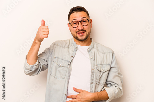 Young hispanic man isolated on white background touches tummy, smiles gently, eating and satisfaction concept. © Asier