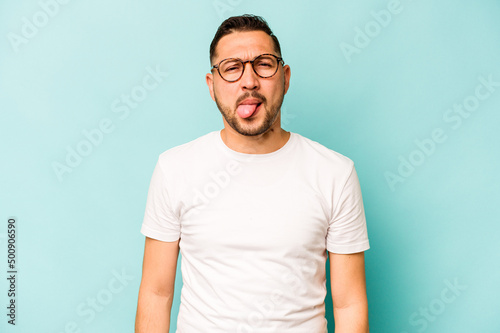 Young hispanic man isolated on blue background funny and friendly sticking out tongue.