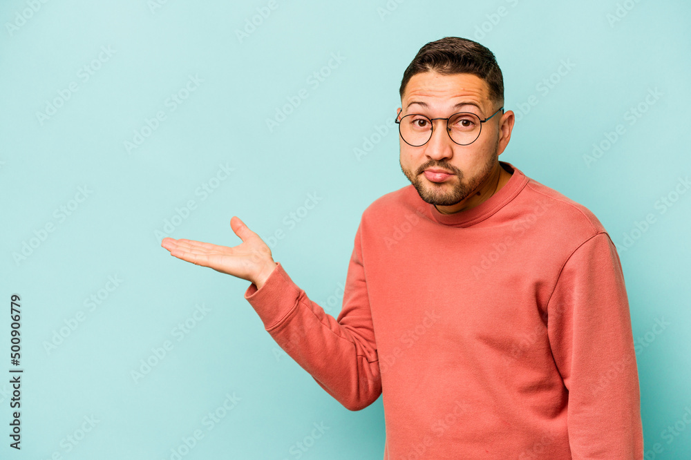 Young hispanic man isolated on blue background showing a copy space on a palm and holding another hand on waist. Young hispanic man isolated on blue background showing a copy space on a palm and holding another hand on waist.