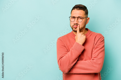 Young hispanic man isolated on blue background looking sideways with doubtful and skeptical expression.
