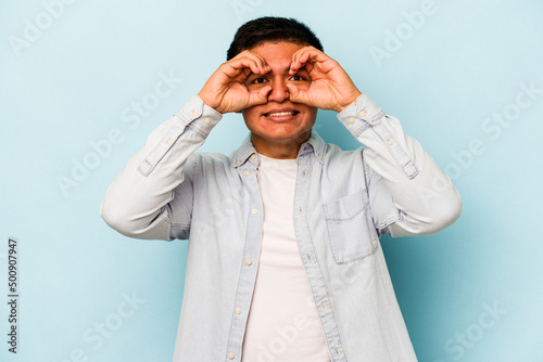 Young hispanic man isolated on blue background showing okay sign over eyes