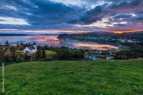 Panorama of the Isle of Skye in the evening. Small town of Uig in Scotland in summer. Evening mood at sunset. Church and other buildings on a hill in a meadow Cloudy sky. Illuminated port.