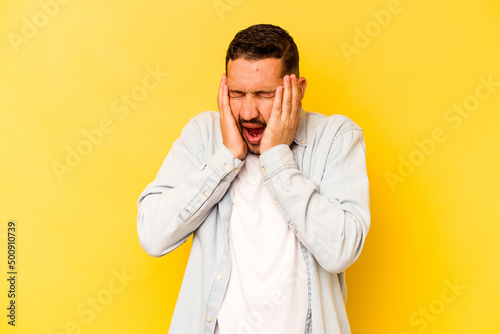 Young hispanic man isolated on yellow background covering ears with hands trying not to hear too loud sound.
