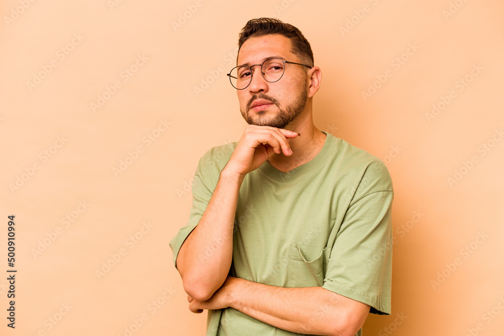 Young hispanic man isolated on beige background suspicious, uncertain, examining you. Young hispanic man isolated on beige background suspicious, uncertain, examining you.