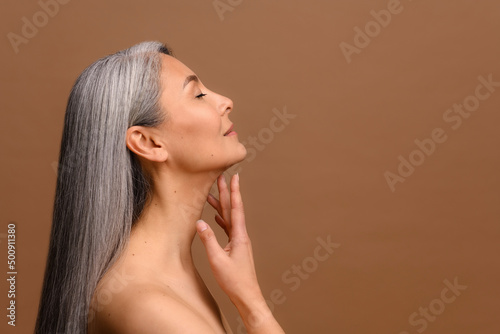 Side view at enchanting topless middle aged Asian woman isolated on brown background, charming korean lady with grey hair stands in profile and touching chin gently. Skin care and self love concept