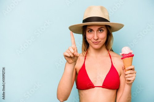 Young caucasian woman wearing a bikini and holding an ice cream isolated on blue background showing number one with finger.