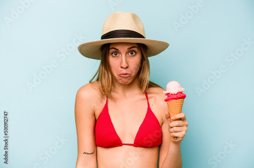 Young caucasian woman wearing a bikini and holding an ice cream isolated on blue background shrugs shoulders and open eyes confused.