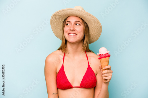Young caucasian woman wearing a bikini and holding an ice cream isolated on blue background dreaming of achieving goals and purposes