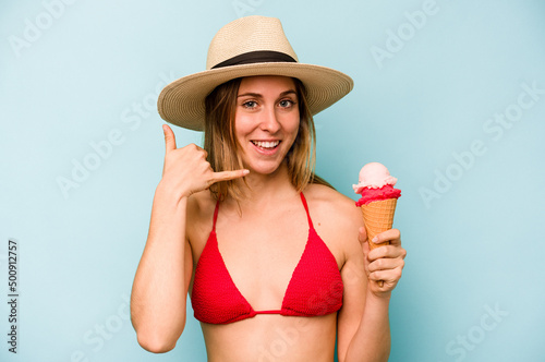 Young caucasian woman wearing a bikini and holding an ice cream isolated on blue background showing a mobile phone call gesture with fingers.