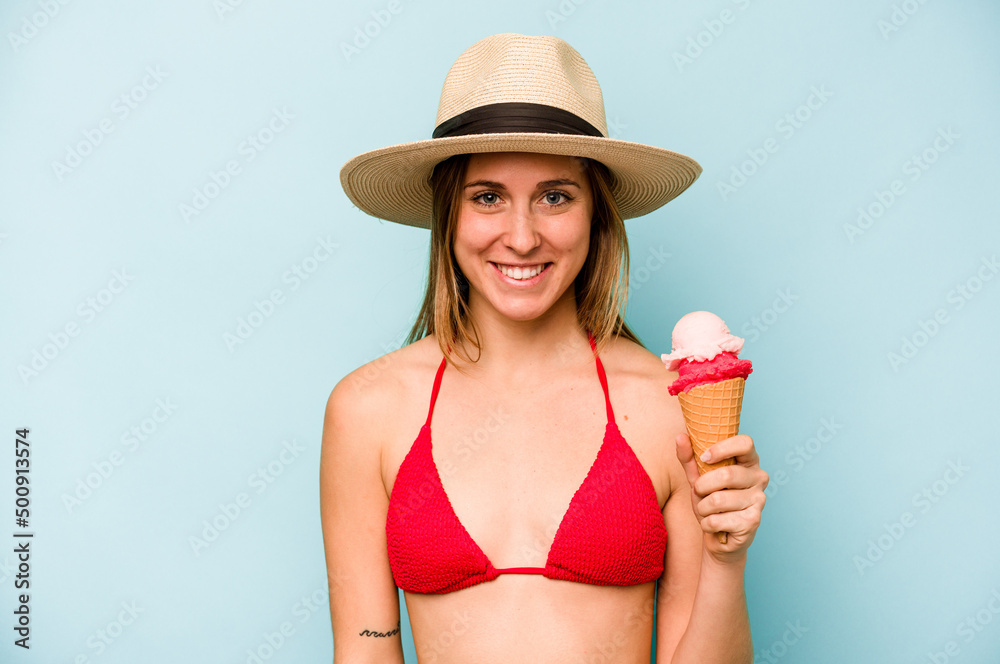 Young caucasian woman wearing a bikini and holding an ice cream isolated on blue background happy, smiling and cheerful.