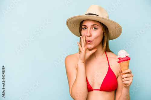 Young caucasian woman wearing a bikini and holding an ice cream isolated on blue background is saying a secret hot braking news and looking aside