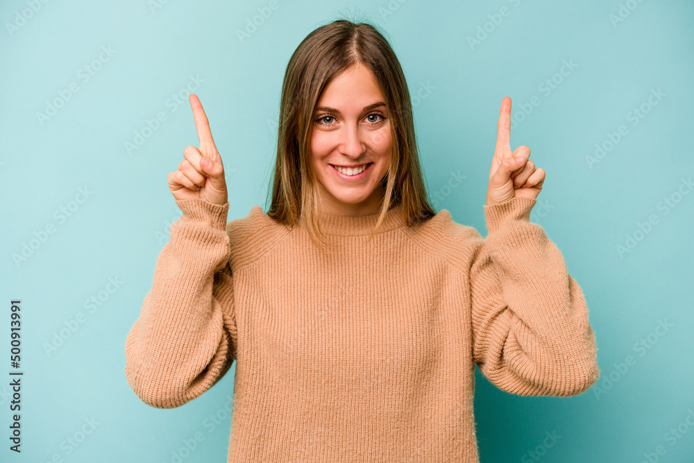 Young caucasian woman isolated on blue background indicates with both fore fingers up showing a blank space. Young caucasian woman isolated on blue background indicates with both fore fingers up showing a blank space.