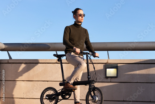 Young man on sunglasses riding an e-bike around the city park under blue sky with sunset light on his face