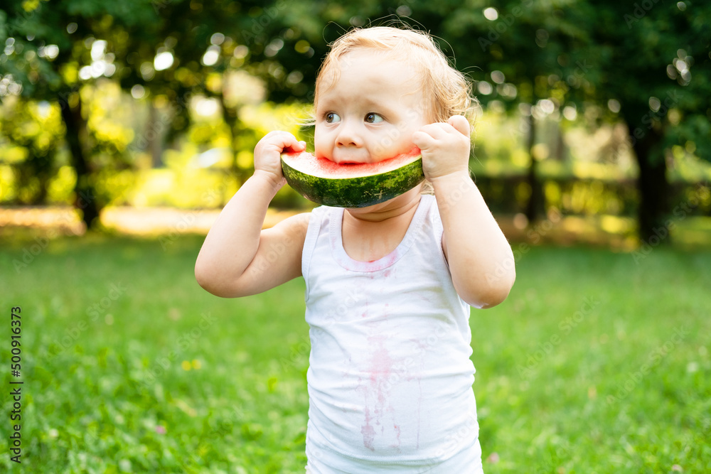 funny smiling kid boy in white bodysuit eating watermelon at green lawn funny smiling kid boy in white bodysuit eating watermelon at green lawn