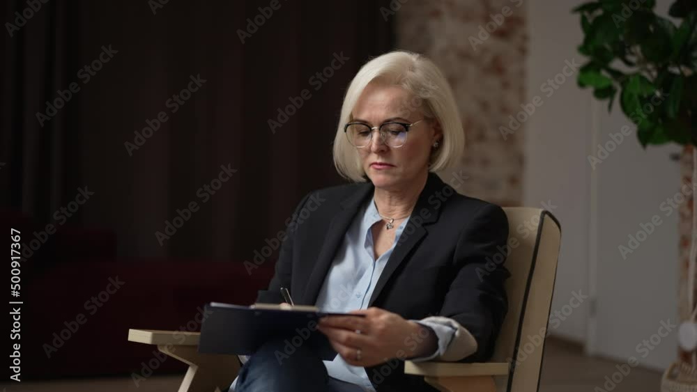 Woman psychotherapist is writing on clipboard and speaking during psychotherapy in office spbas. 4k Portrait of beautiful European female makes notes and speaks with smile, gestures with hand and sits Woman psychotherapist is writing on clipboard and speaking during psychotherapy in office spbas. 4k Portrait of beautiful European female makes notes and speaks with smile, gestures with hand and sits