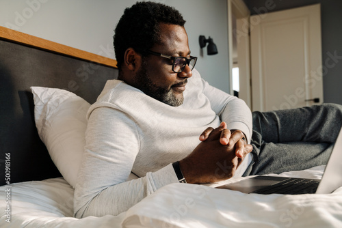 Black man wearing eyeglasses using laptop while resting in bed