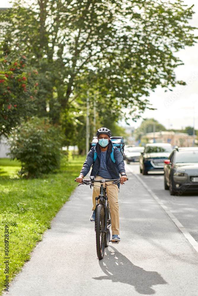 Fototapeta premium food shipping, health and people concept - delivery man in bike helmet and protective medical mask with thermal insulated bag and smatphone riding bicycle on city street
