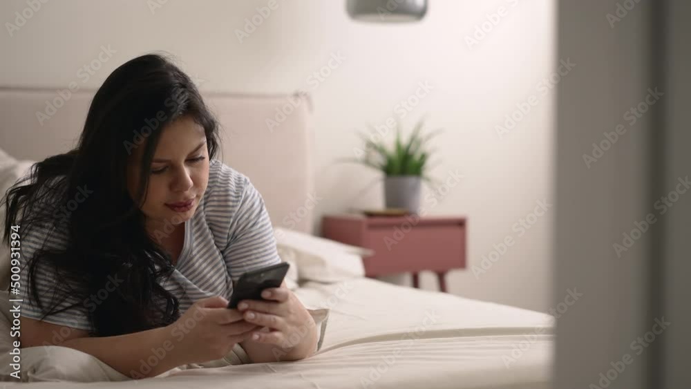 Young overweight woman holding smartphone in hand and lying on bed in home bedroom spbd. 4k Close view of oversized European female uses phone and looks at screen, searches in internet or types Young overweight woman holding smartphone in hand and lying on bed in home bedroom spbd. 4k Close view of oversized European female uses phone and looks at screen, searches in internet or types