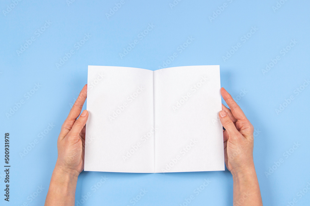 Obraz premium Woman hands holding folded blank paper sheet, empty booklet, double-pages book, magazine on light blue background. Mock up template. Top view