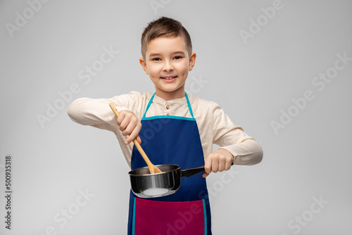 culinary and profession concept - happy smiling little boy in apron with saucepan and wooden spoon cooking food over grey background