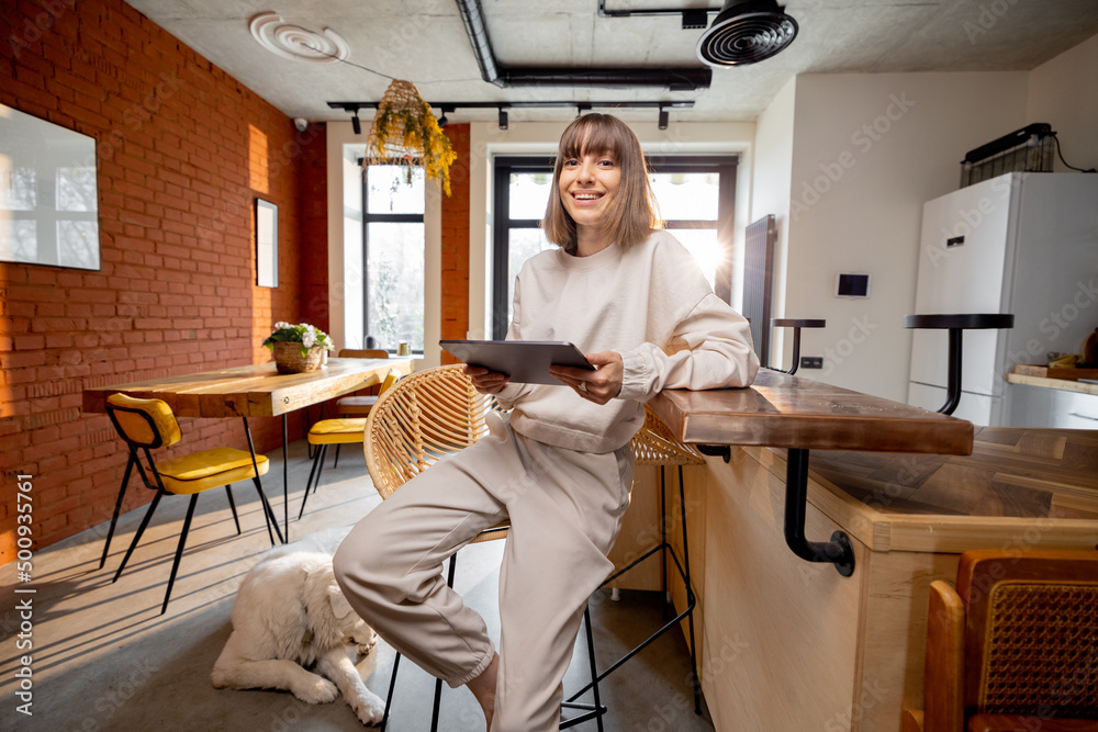 Young pretty woman in domestic wear sitting with a digital tablet on bar chair at modern studio apartment in loft style. Concept of work from home Young pretty woman in domestic wear sitting with a digital tablet on bar chair at modern studio apartment in loft style. Concept of work from home