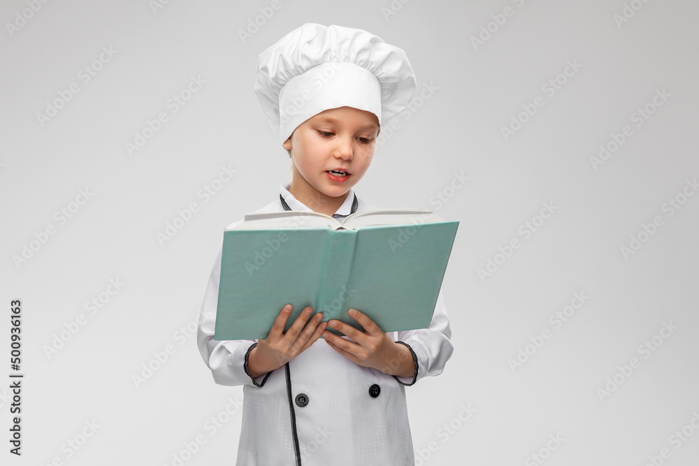 cooking, culinary and profession concept - happy smiling little girl in chef's toque and jacket reading cook book over grey background cooking, culinary and profession concept - happy smiling little girl in chef's toque and jacket reading cook book over grey background