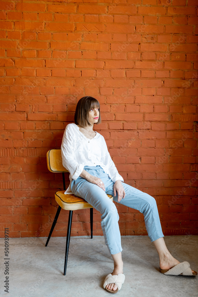 Full length portrait of a young woman dressed casually sits relaxed on chair on brick wall background at home