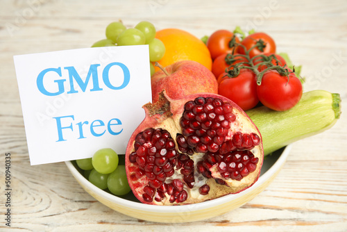 Fresh fruits, vegetables and card with text GMO Free in bowl on white wooden table