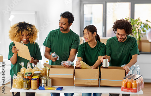charity, donation and volunteering concept - international group of happy smiling volunteers packing food in boxes according to list on clipboard at distribution or refugee assistance center