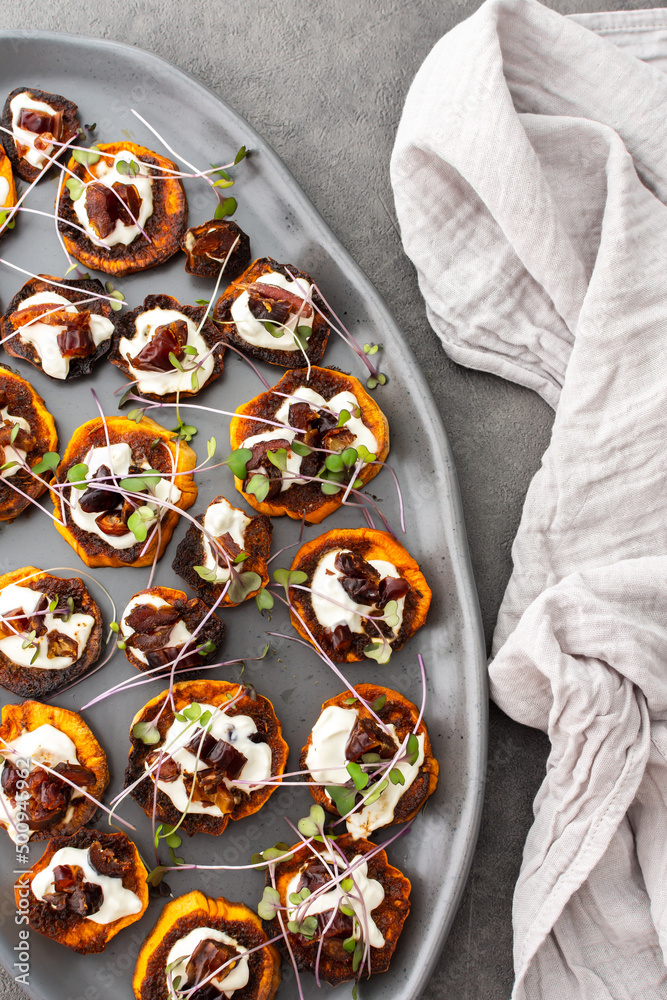 Oven baked slices of sweet orange sweet potato with yogurt and dates, sprinkled with microgreens, sweet potato on a gray oval ceramic dish, top view