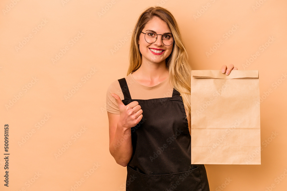 Restaurant waiter caucasian woman holding a take away bag isolated on beige background smiling and raising thumb up Restaurant waiter caucasian woman holding a take away bag isolated on beige background smiling and raising thumb up