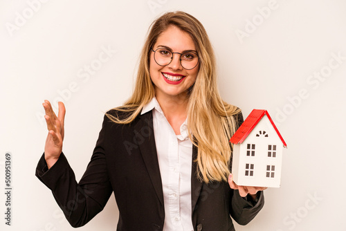 Young business caucasian woman holding a toy house isolated on white background receiving a pleasant surprise, excited and raising hands.