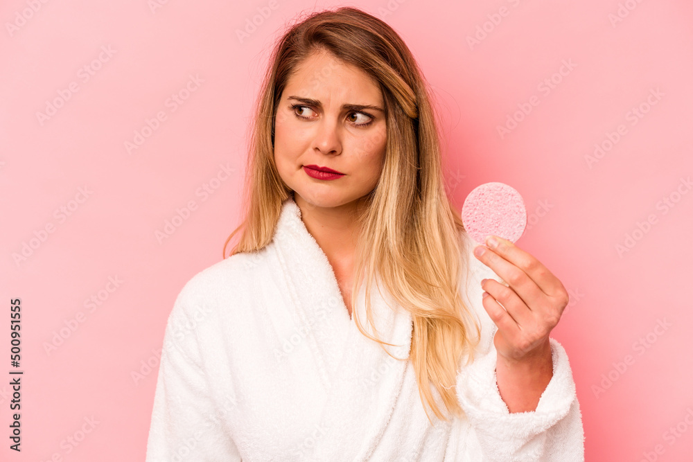 Young caucasian woman holding facial sponge isolated on pink background confused, feels doubtful and unsure. Young caucasian woman holding facial sponge isolated on pink background confused, feels doubtful and unsure.