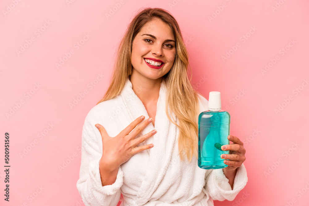 Young caucasian woman holding mouthwash wearing bathrobe isolated on pink background laughs out loudly keeping hand on chest. Young caucasian woman holding mouthwash wearing bathrobe isolated on pink background laughs out loudly keeping hand on chest.