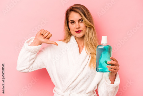 Young caucasian woman holding mouthwash wearing bathrobe isolated on pink background feels proud and self confident, example to follow.