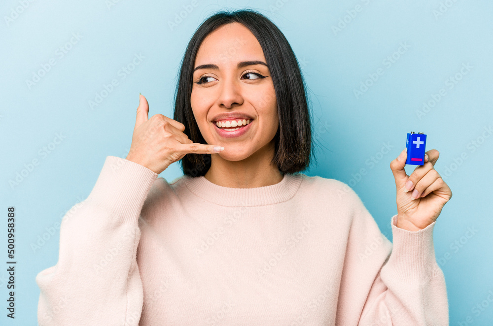 Young hispanic woman holding batteries isolated on blue background showing a mobile phone call gesture with fingers. Young hispanic woman holding batteries isolated on blue background showing a mobile phone call gesture with fingers.