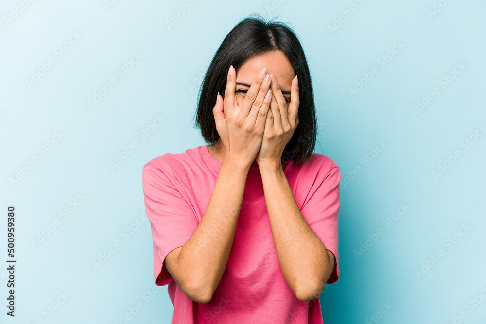 Young hispanic woman isolated on blue background blink at the camera through fingers, embarrassed covering face. Young hispanic woman isolated on blue background blink at the camera through fingers, embarrassed covering face.
