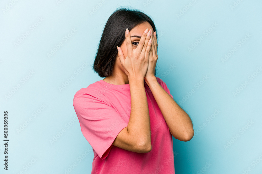 Young hispanic woman isolated on blue background blink through fingers frightened and nervous.