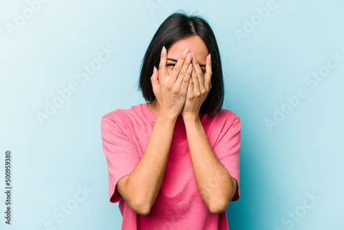 Young hispanic woman isolated on blue background blink at the camera through fingers, embarrassed covering face. © Asier