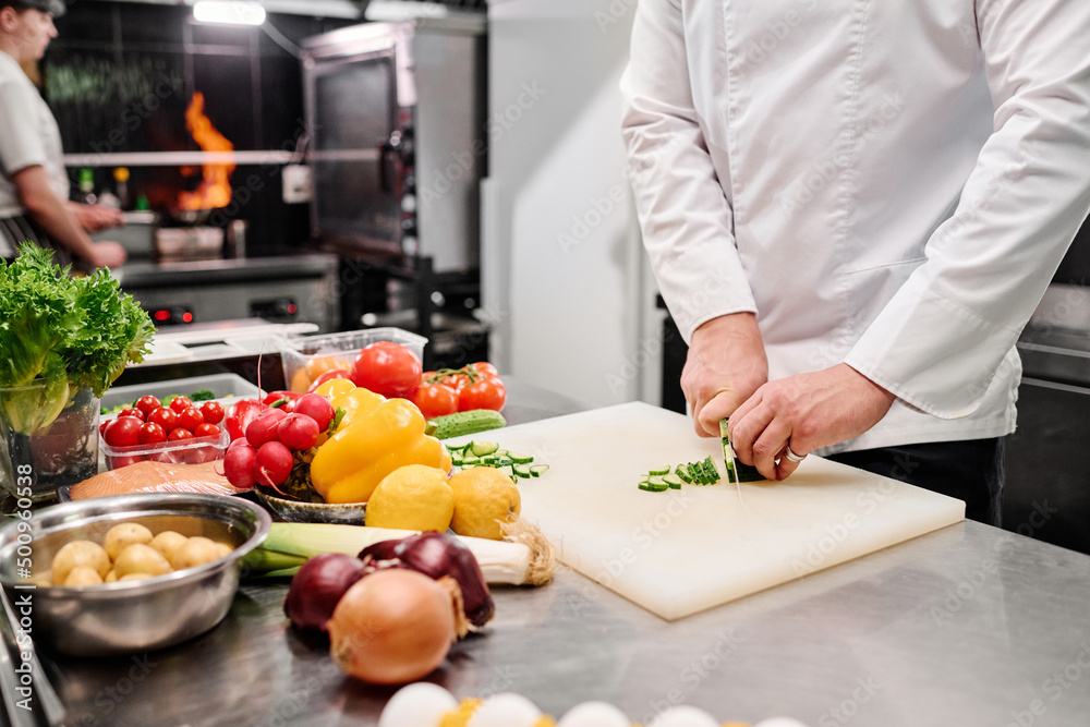 Close-up of cook in uniform cutting fresh vegetable on cutting board at table during work in kitchen Close-up of cook in uniform cutting fresh vegetable on cutting board at table during work in kitchen