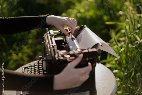 hands loading paper into a Retro typewriter with blank sheet. Dream writer's workplace in green wheat field. Inspiration, Creative freedom and remote work concept