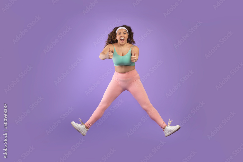 Satisfied excited young european plus size woman in sportswear jump, freeze in air, show thumbs up
