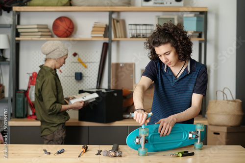 Contemporary teenager with handtool fixing wheels of skateboard by wooden table or workbench against his grandmother with magazine