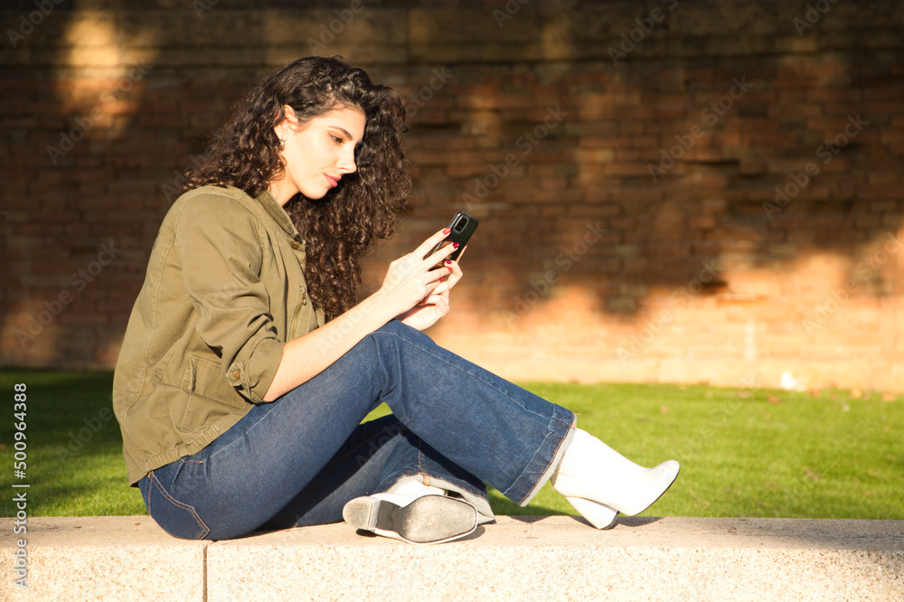 Beautiful young latina woman with curly brown hair and dressed in casual clothes sitting on a grey wall checking her mobile phone, social media, shopping and email. Technology concept.
