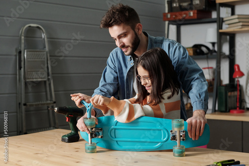 Cuadro en lienzo Cute smiling girl with handtool fixing wheels of skateboard on wooden table in g