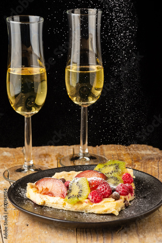 Close-up of piece of fruit cheesecake with falling sugar, on table with two glasses of champagne, black background, vertical