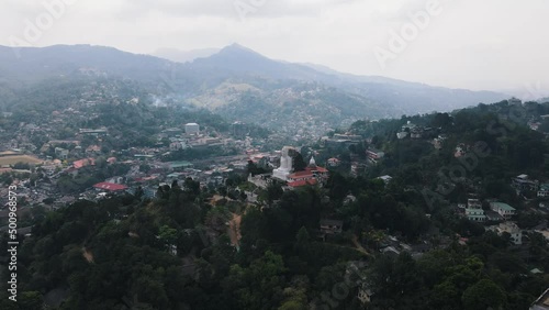 Large white Bahirawakanda Vihara Buddha Statue on one of the green hills in the mountainous landscape of Sri Lanka with the large city of Kandy in the valley on a cloudy day. Wide drone panning shot photo