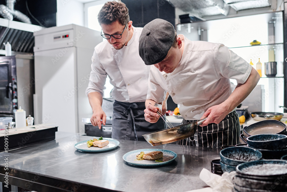 Cook in uniform putting garnish on plate from pan with chef standing behind him and controlling the process Cook in uniform putting garnish on plate from pan with chef standing behind him and controlling the process