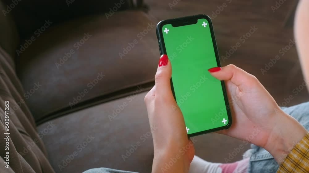 Young woman sitting on sofa at home and uses smartphone with green screen Mock-up, scroll motion. 4k	
